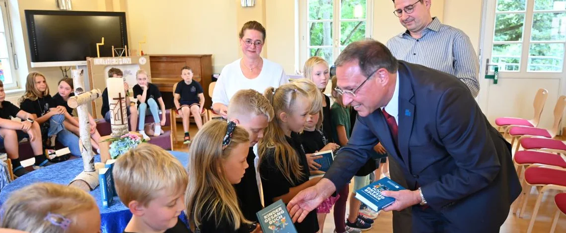 Stiftungspfarrer Dr. Ekkehard Steinhäuser von der Evangelischen Schulstiftung in Mitteldeutschland übergibt in der Evangelischen Grundschule Zerbst die neuen Kindergesangbücher an Schülerinnen und Schüler der ersten Klasse. Im Hintergrund Oberkirchen